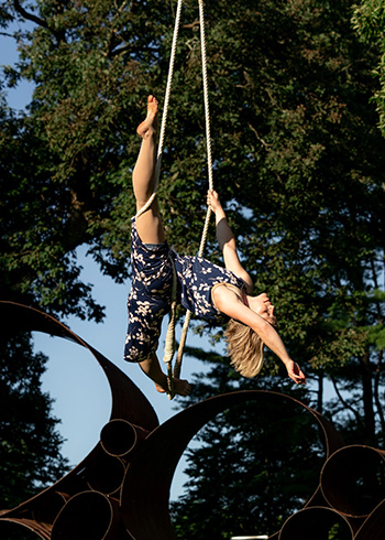 A woman, Alissa Feller, is suspended in midair for an outdoor trapeze performance.