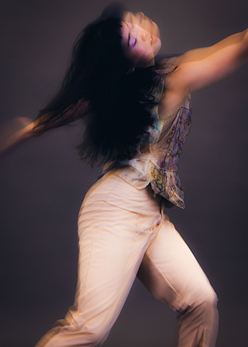 An intentionally artistically blurry photo shows a woman, Cassie Wang, dancing, she is turned to one side with her knees slightly bent and arms stretched out forming a diagonal.