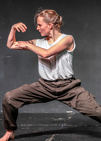 A woman, Savannah Dunn, dances onstage, she faces sideways and crouches in a lunge and touches one bicep with her opposite hand.