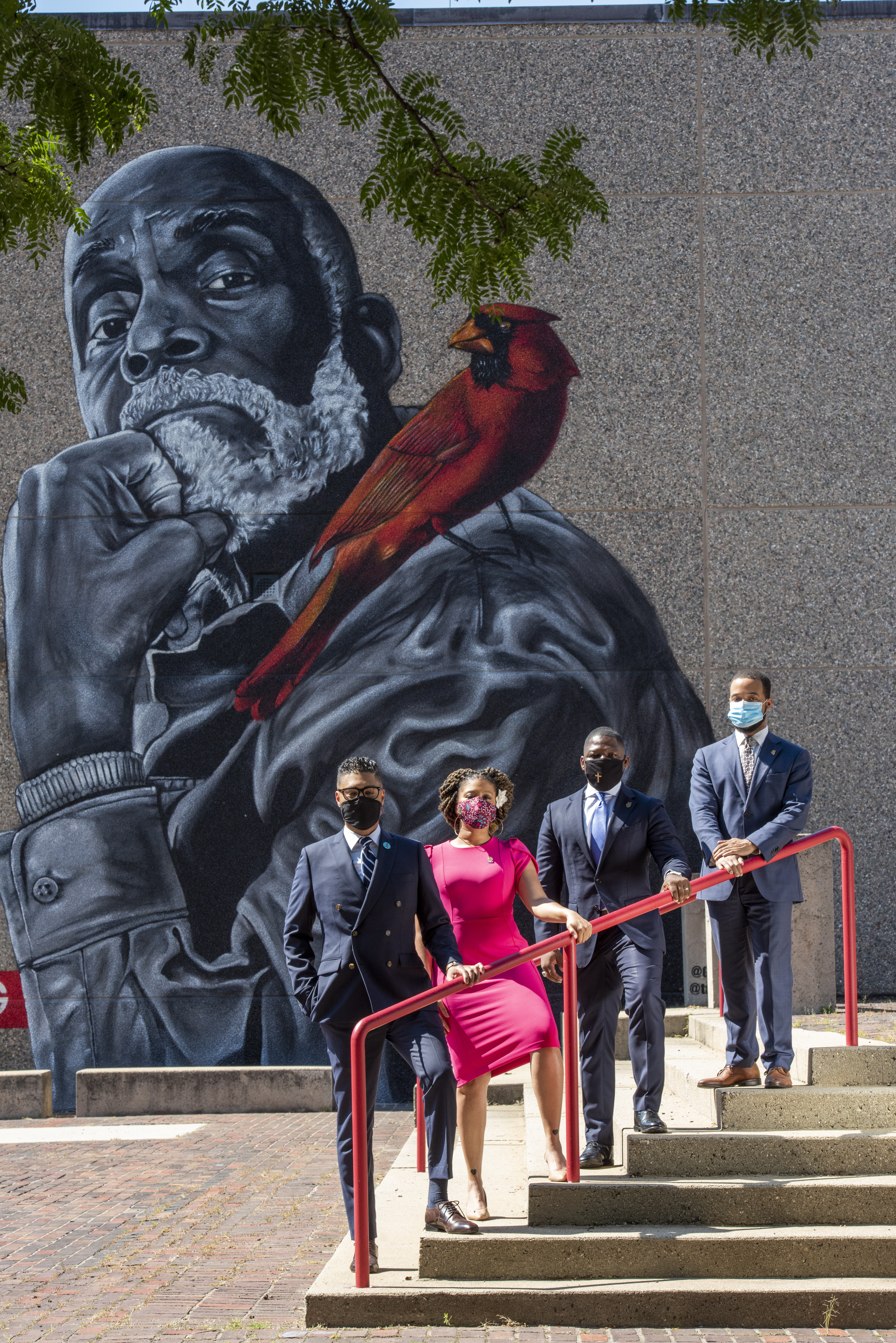 Imari Paris Jeffries, Sheena Collier, Willie Brodick and Segun Idowu standing in a row on a set of concrete stairs, outside with masks on. Behind them is a concrete wall witha. large mural of Mel King with a cardinal on his shoulder. Branches and leaves of a tree peak into the top of the frame. 