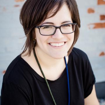 A headshot photo of a woman, she is smiling, wearing glasses, and has shoulder length brown hair