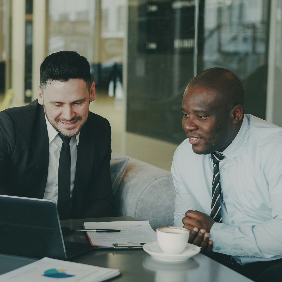 Two men look at a laptop with documents in front of them