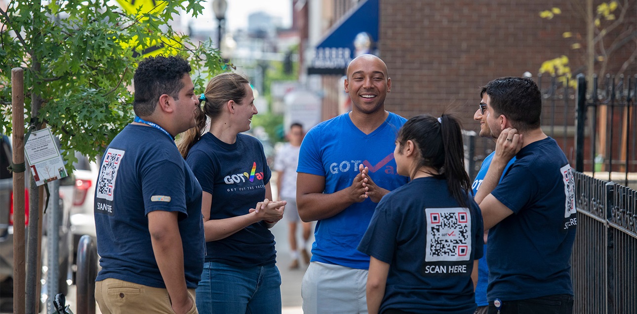 A group of people is gathered on a sidewalk outside a building. They are all wearing shirts with GoToVote logo on the front and a QR code on the back.