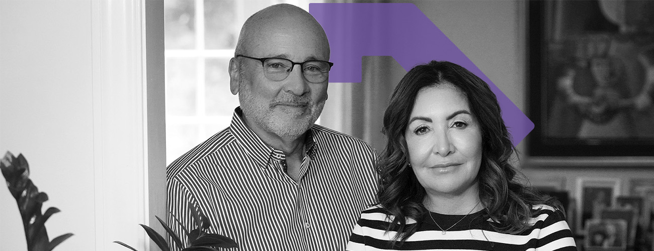 A black and white photo of two donors standing in a home posing for a photo. Edited into the photo layered behind the subject is the purple TBF arrow icon