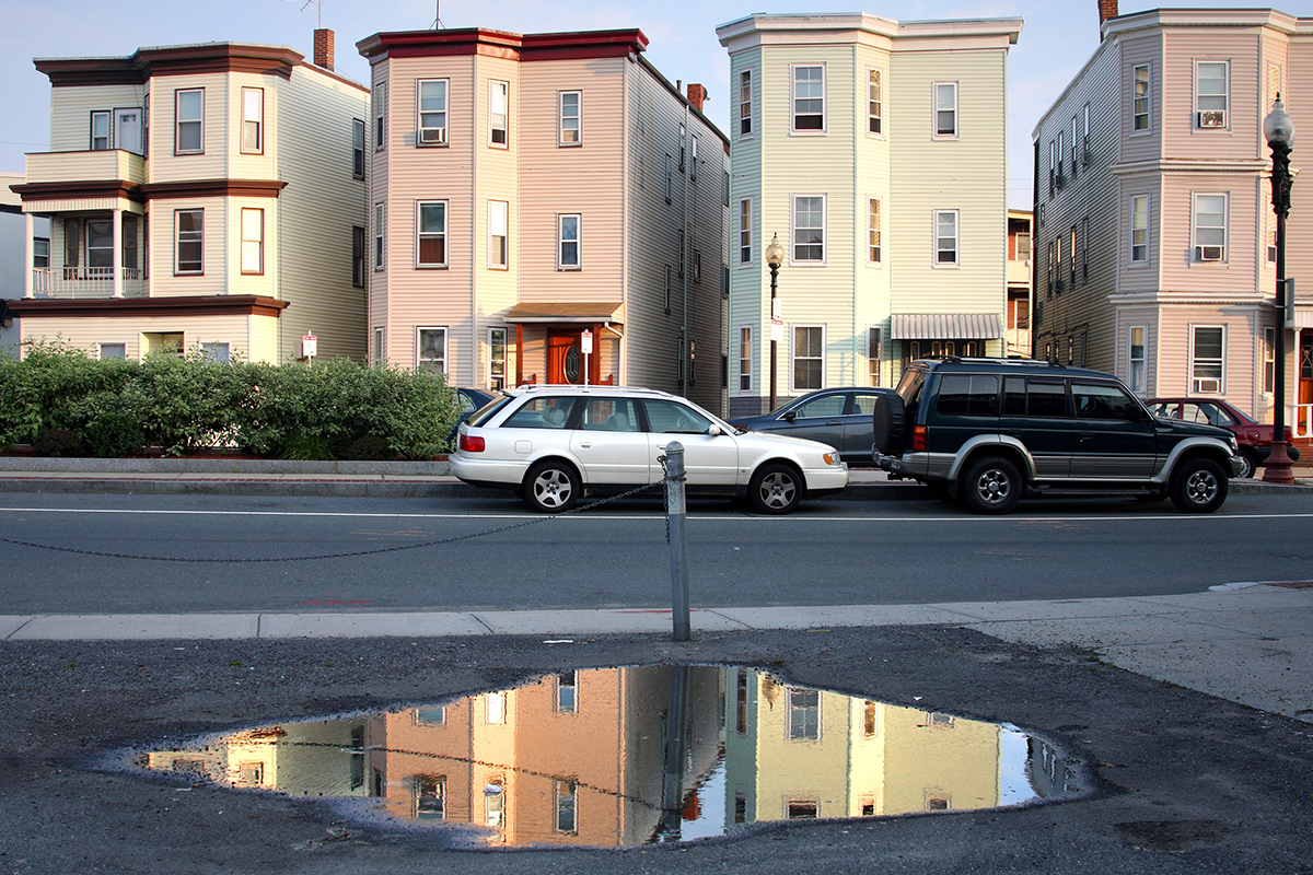 A photo of a street shows a few houses and a parking lot with a large puddle