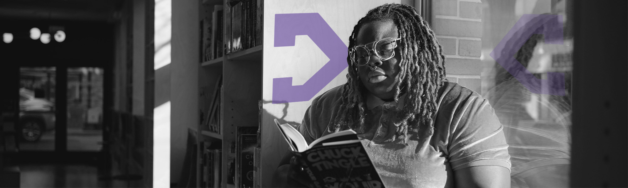 A Black woman with glasses and long locs sits by a window reading with a TBF logo reflected behind her