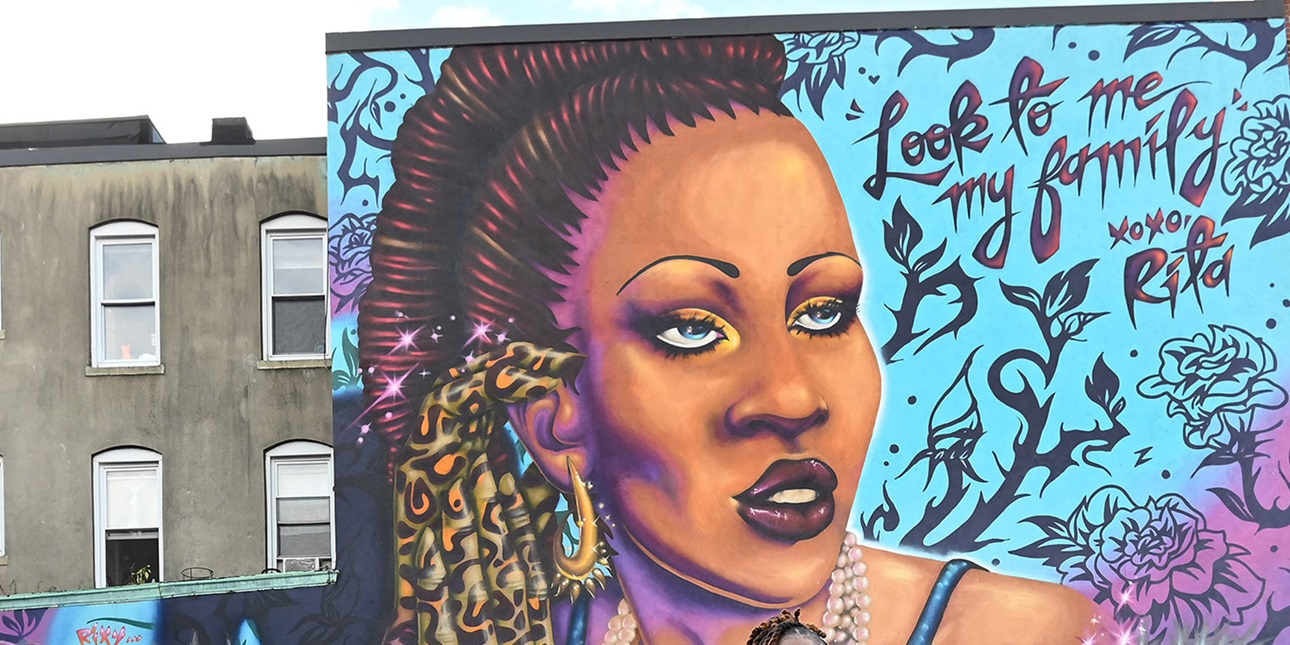 A colorful mural on the side of a brick building depicts a trans woman. Text reads "look to me my family, xoxo Rita."
