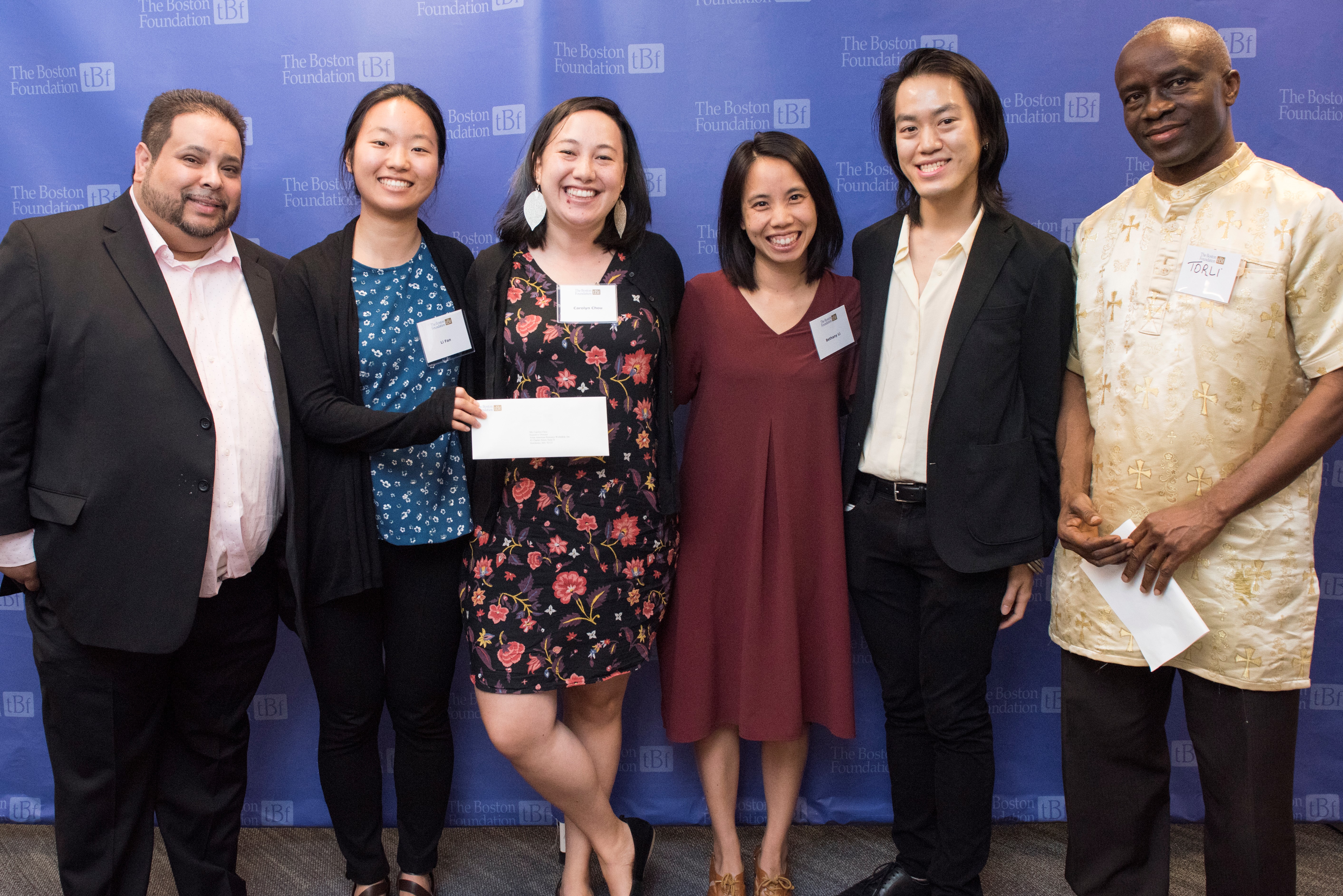 A group of six men and women stand in front of a blue TBF backdrop and pose for a photo, one woman is holding a piece of paper