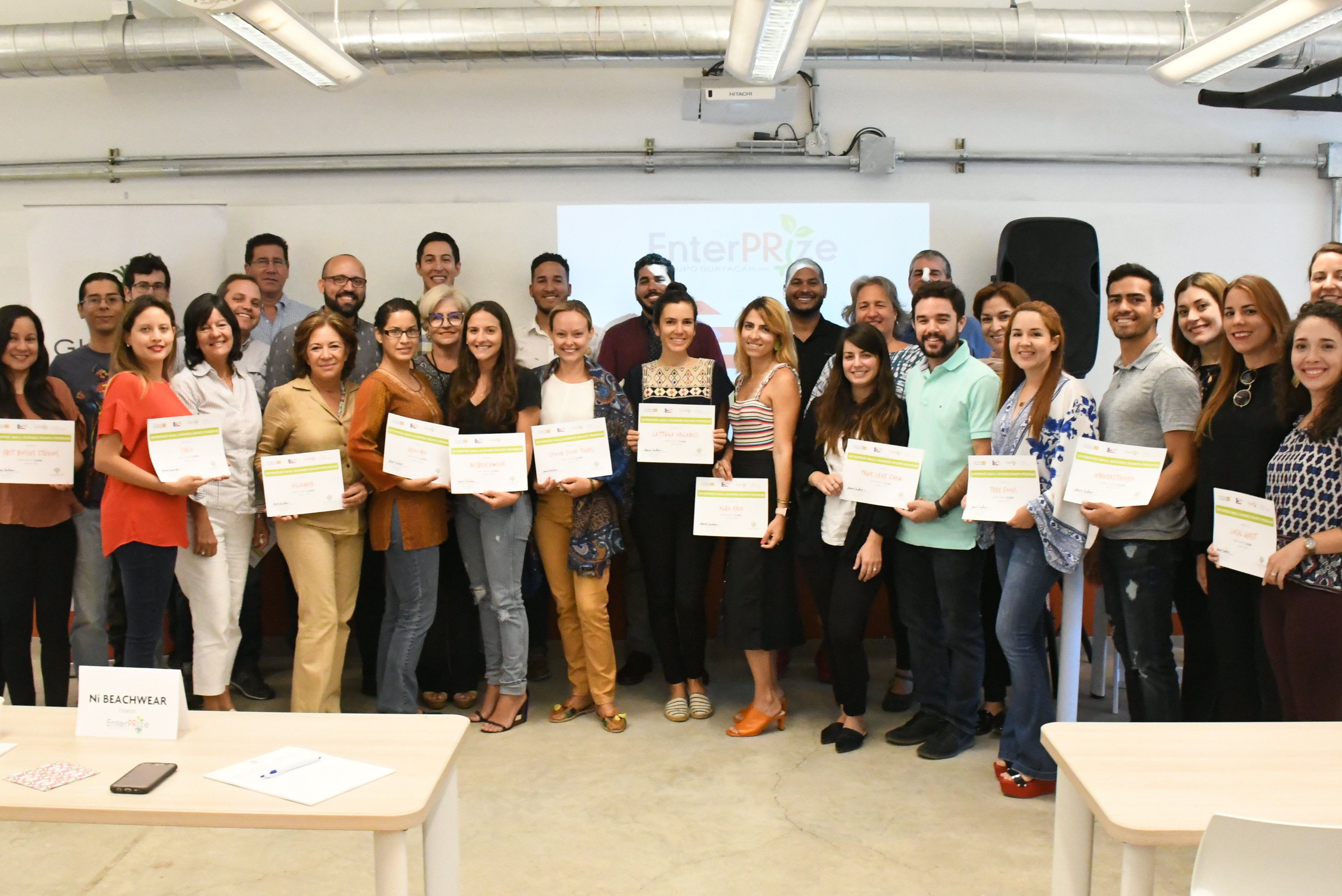 A large group of people stand at the front of a classroom. They are all holding paper certificates and posing for a photo
