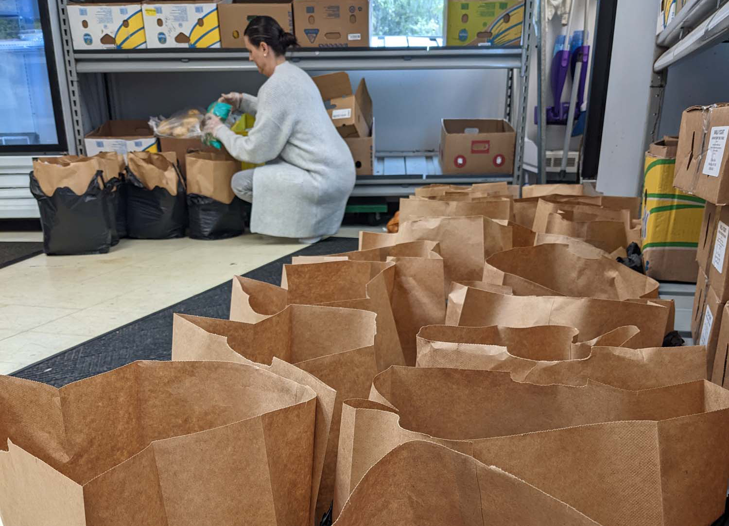 A photo taken in a food pantry, a woman crouches down and packs food into brown bags