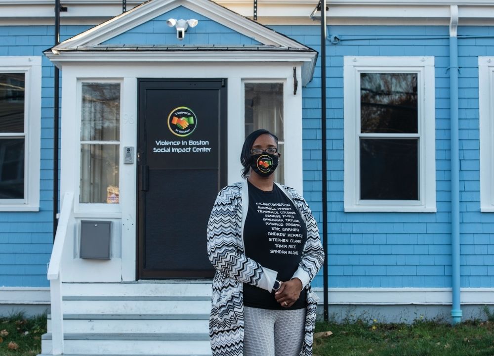 Monica Cannon-Grant standing in front of the doorway to her organization, Violence in Boston. The organization's logo is on the black door, and the building has blue siding. Monica is wearing a black t-shirt with white writing on the front, and a black and white cardigan and gray pants. She's wearing a black face mask with the Violence in Boston logo on it.