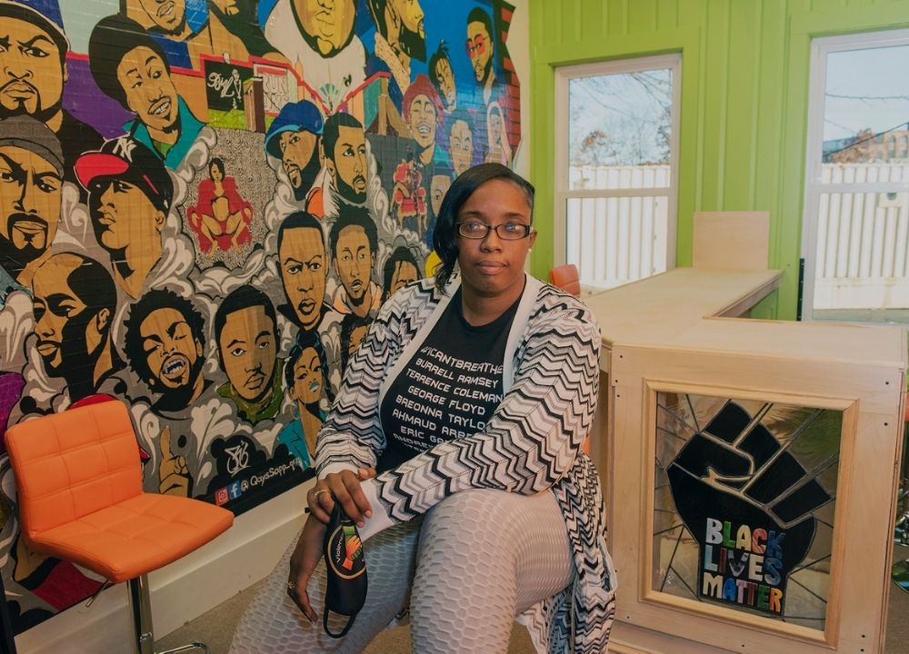 Monica Canon-Grant sitting inside Violence in Boston. Behind her to the left is a colorful mural of the faces of Black and Brown men and women. Behind her to the right is a lime green wall with two windows. To the right is a wooden half-wall with a stained glass window that says "Black Lives Matter" below a black fist.