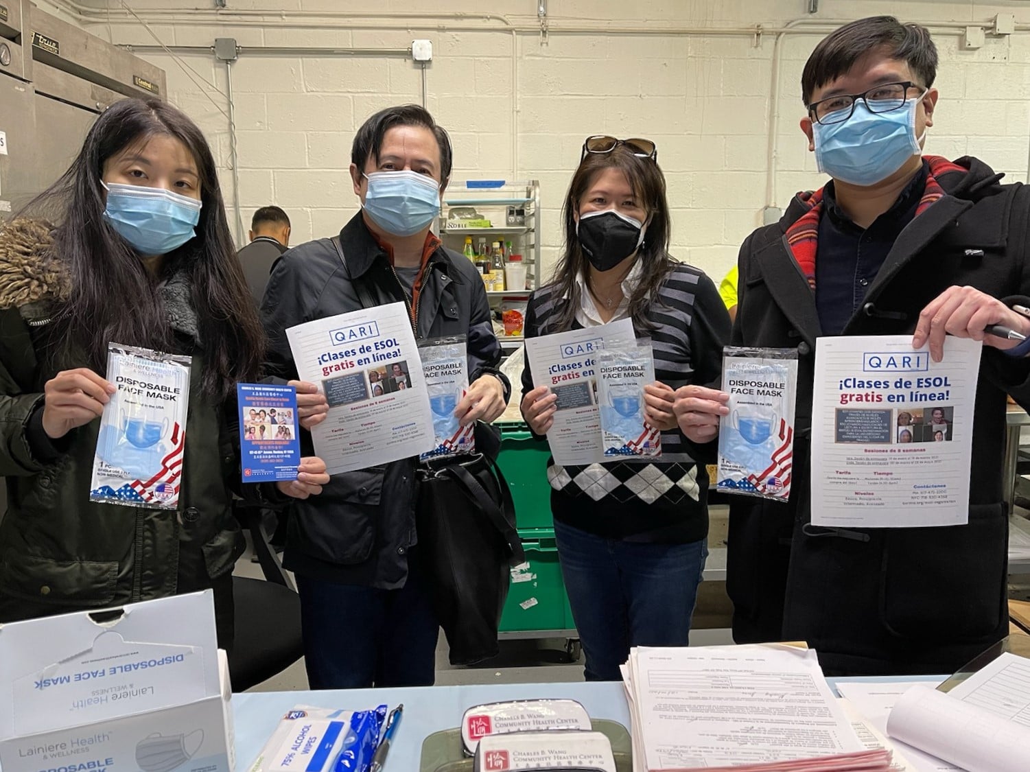 Four QARI volunteers wearing face masks and standing at a table covered in pamphlets about ESOL classes. Each volunteer is holding up a Spanish-language pamplet about ESOL classes.