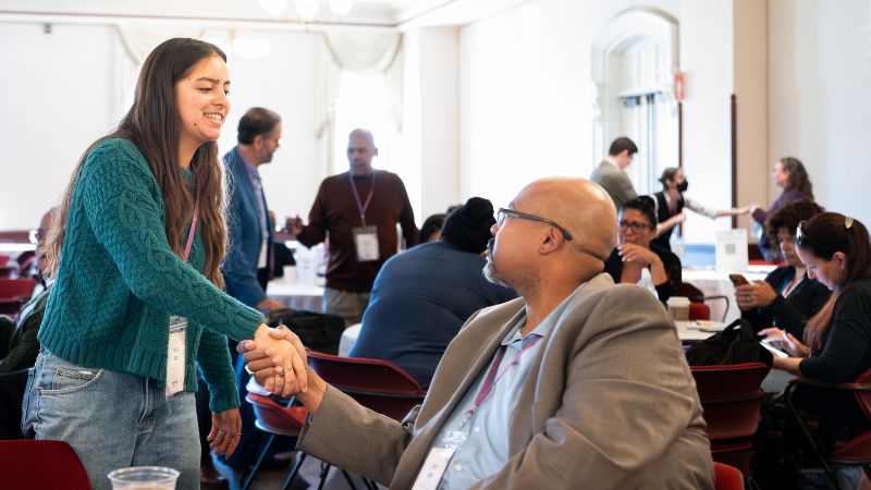 Woman standing shakes hand of man sitting at table at an event.