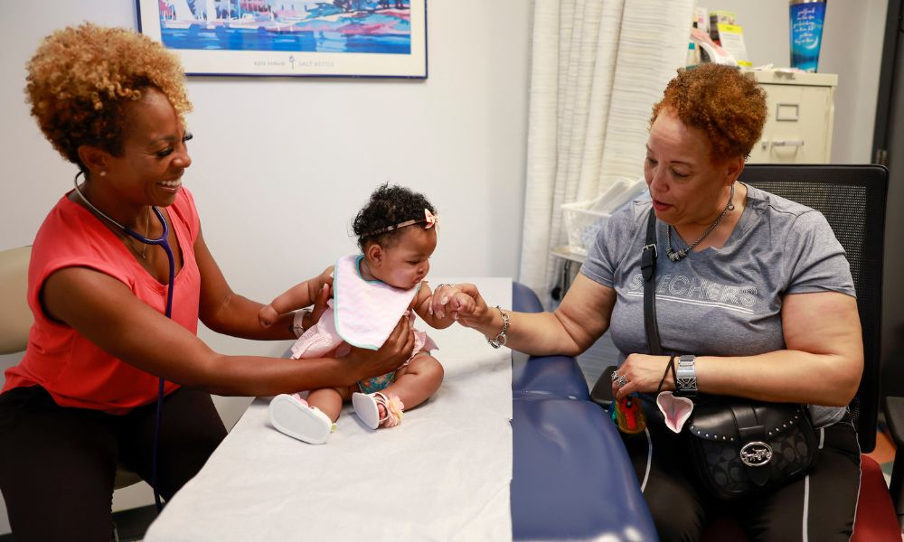 During a visit, a baby girl sits on an exam table, with a practitioner on one side supporting her to sit up and her mom on the other side holding her hand