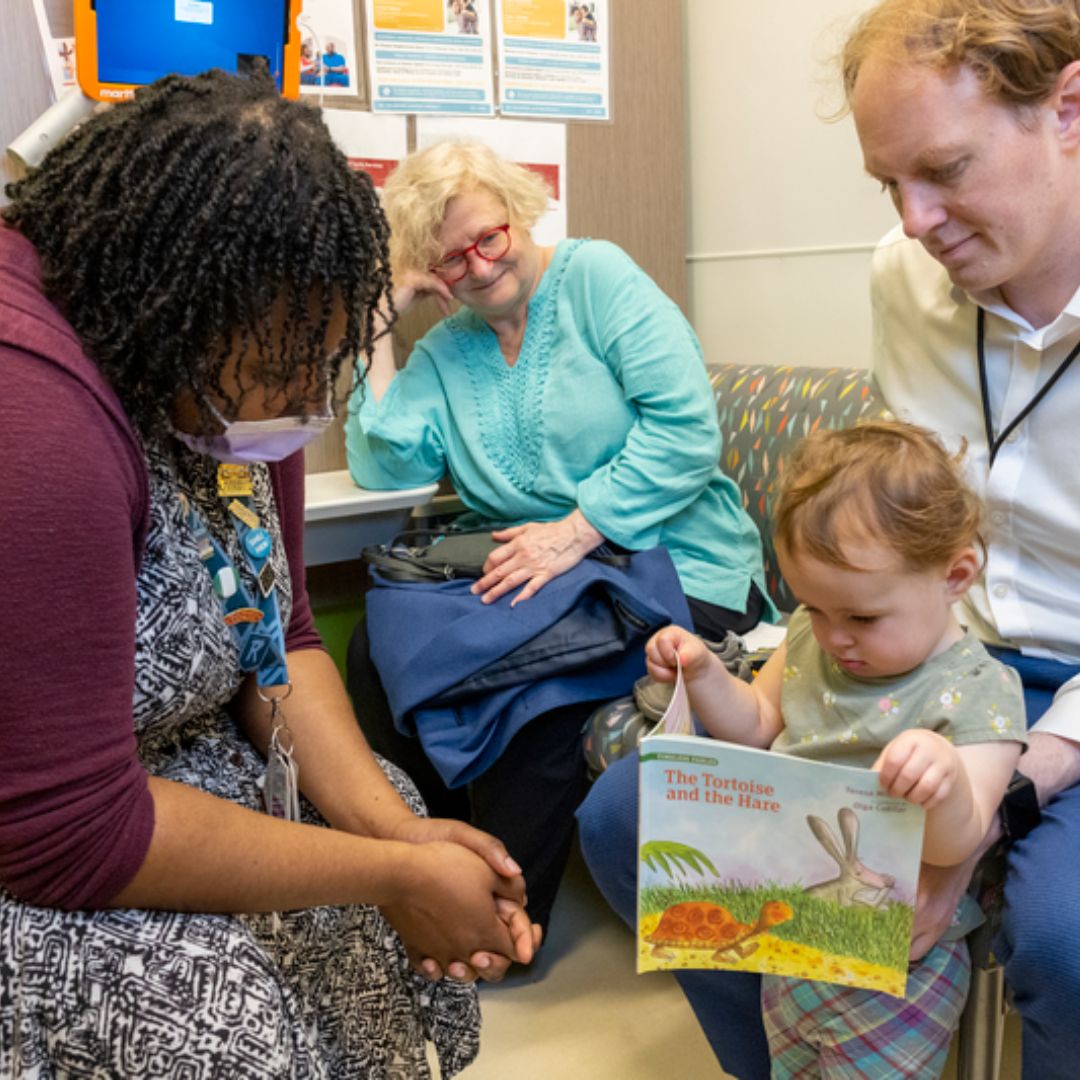 A practitioner leans over to engage with a young child holding a book. The child's parent and grandparent are with him for this visit.