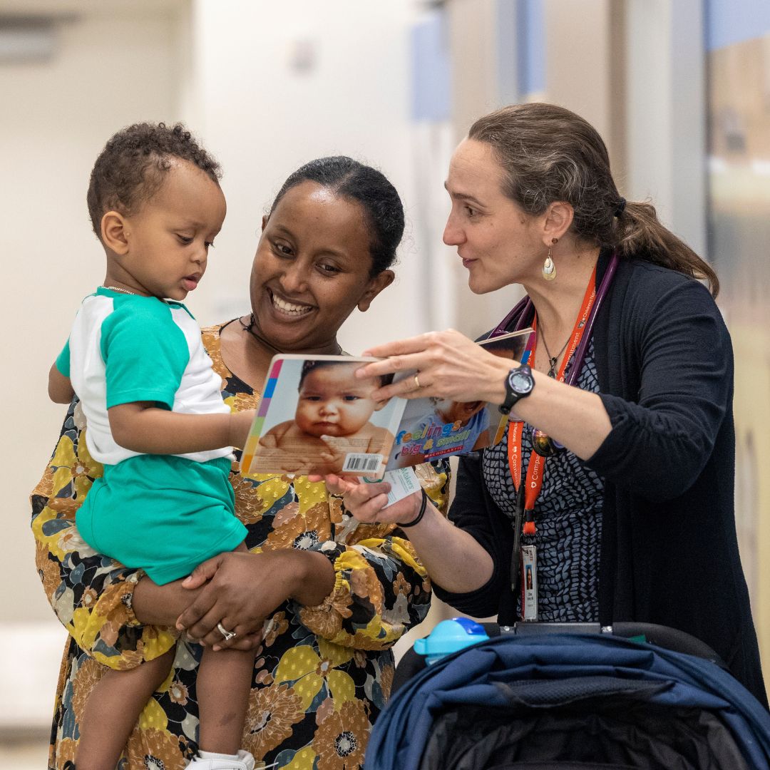 A woman holds a young child while another woman engages them with a colorful children's book. The book features a baby’s face on the cover and has the title “feelings big and small.” The setting is a brightly lit hallway