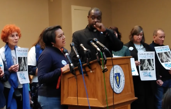 Damaris Velasquez speaking at a podium, people behind her holding signs.