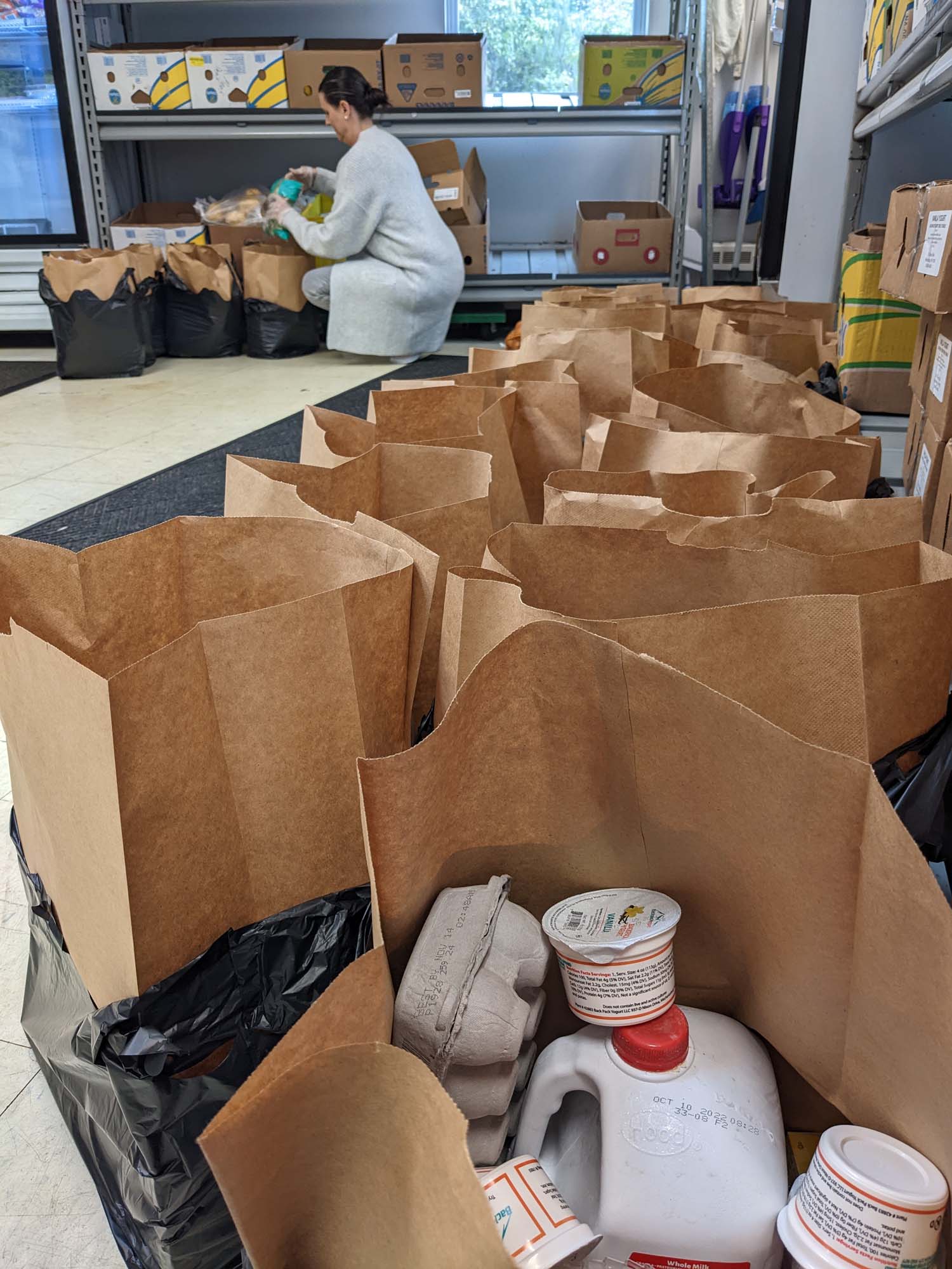 A photo shows many brown bags that are packed with food at a food pantry. In the background a woman fills more bags with food.