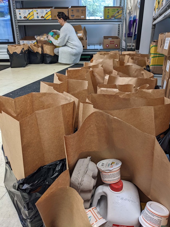 A photo shows many brown bags that are packed with food at a food pantry. In the background a woman fills more bags with food.