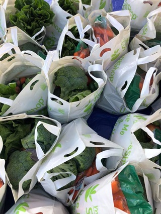 An overhead shot of multiple white bags of groceries filled with produce, broccoli or carrots on the top of each one.