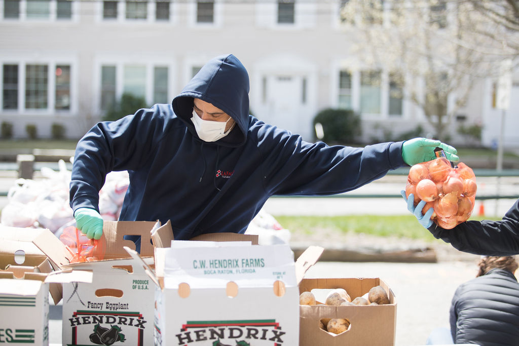 A man wearing a black hoodie and protective mask is taking produce out of boxes and handing it to someone on his left.