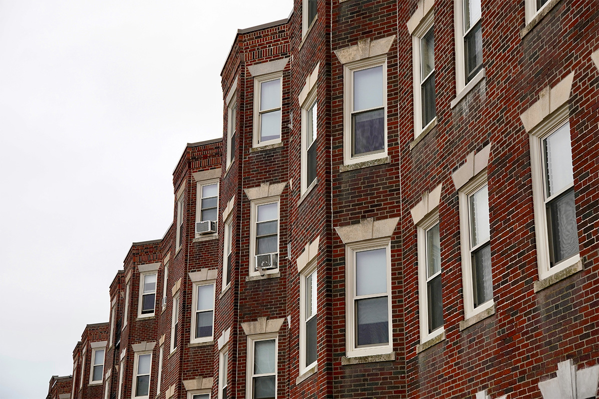 A row of multiple storied brick buildings, photographed upwards so the top stories can be seen along with a cloudy gray sky.