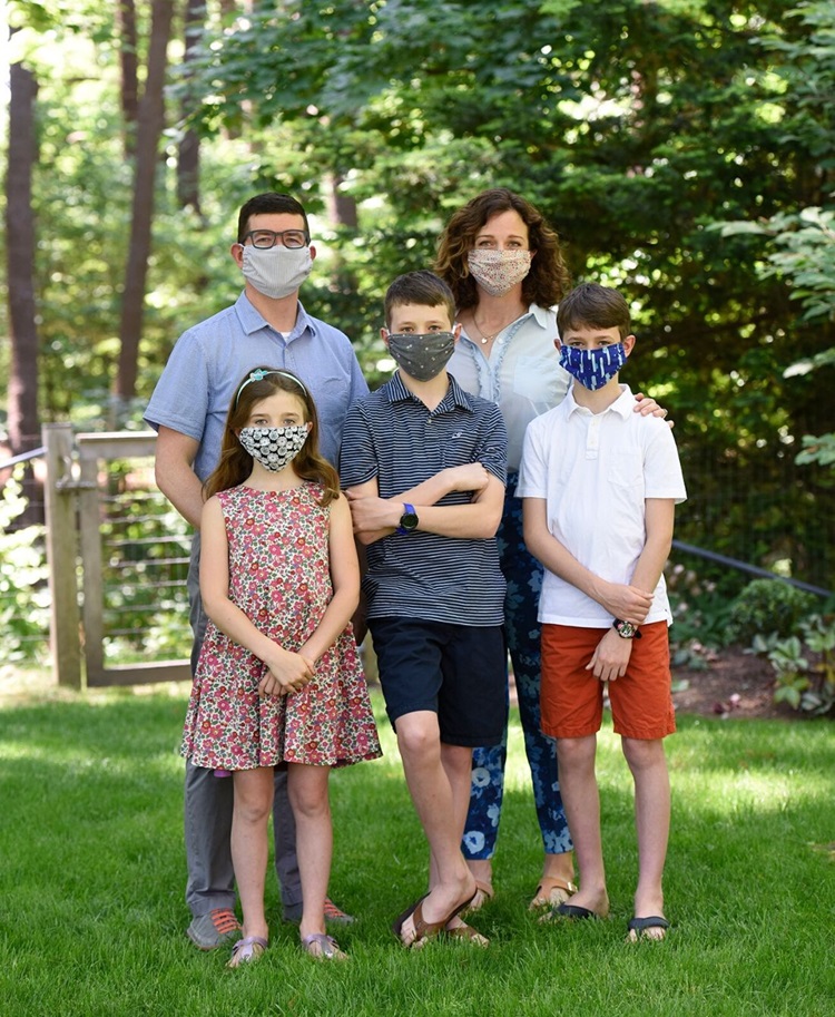 Maggie Schmidt and Ken Danila standing behind their three kids, all standing for a family photo with face masks on, on green grass in front of trees.