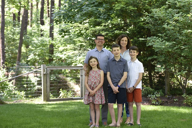 Maggie Schmidt and Ken Danila standing behind their three kids, all standing for a family photo on green grass in front of trees.