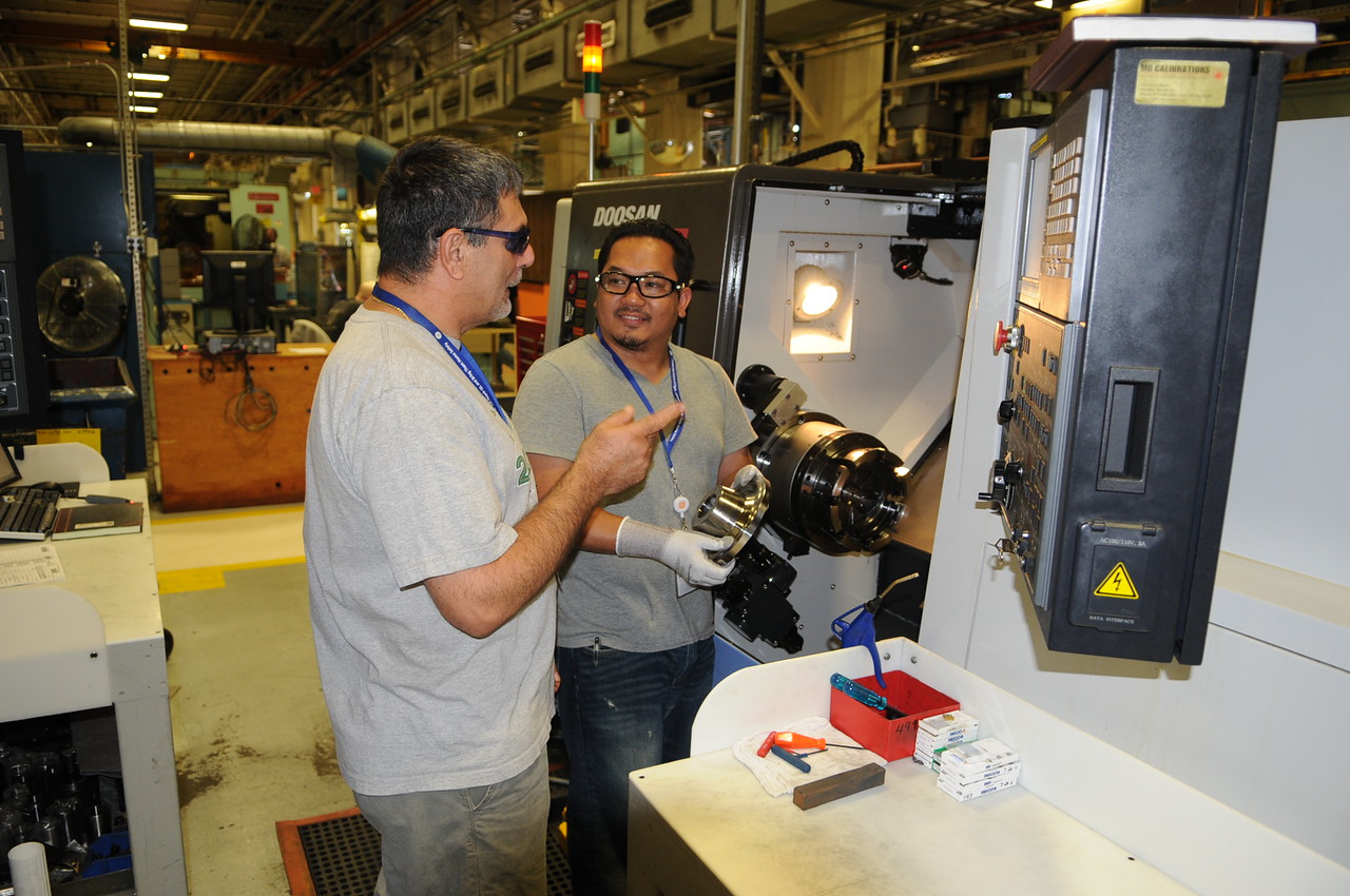 Two men standing wearing protective goggles, talking to each other in front of a machine inside a machine workshop.
