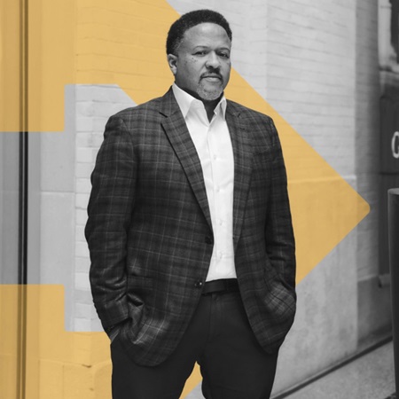 A black-and-white photo of Orlando Watkins, a Black man with short hair and a beard, standing with hands in his suit pockets on a streetscape. A TBF orange arrow graphic is inserted behind him