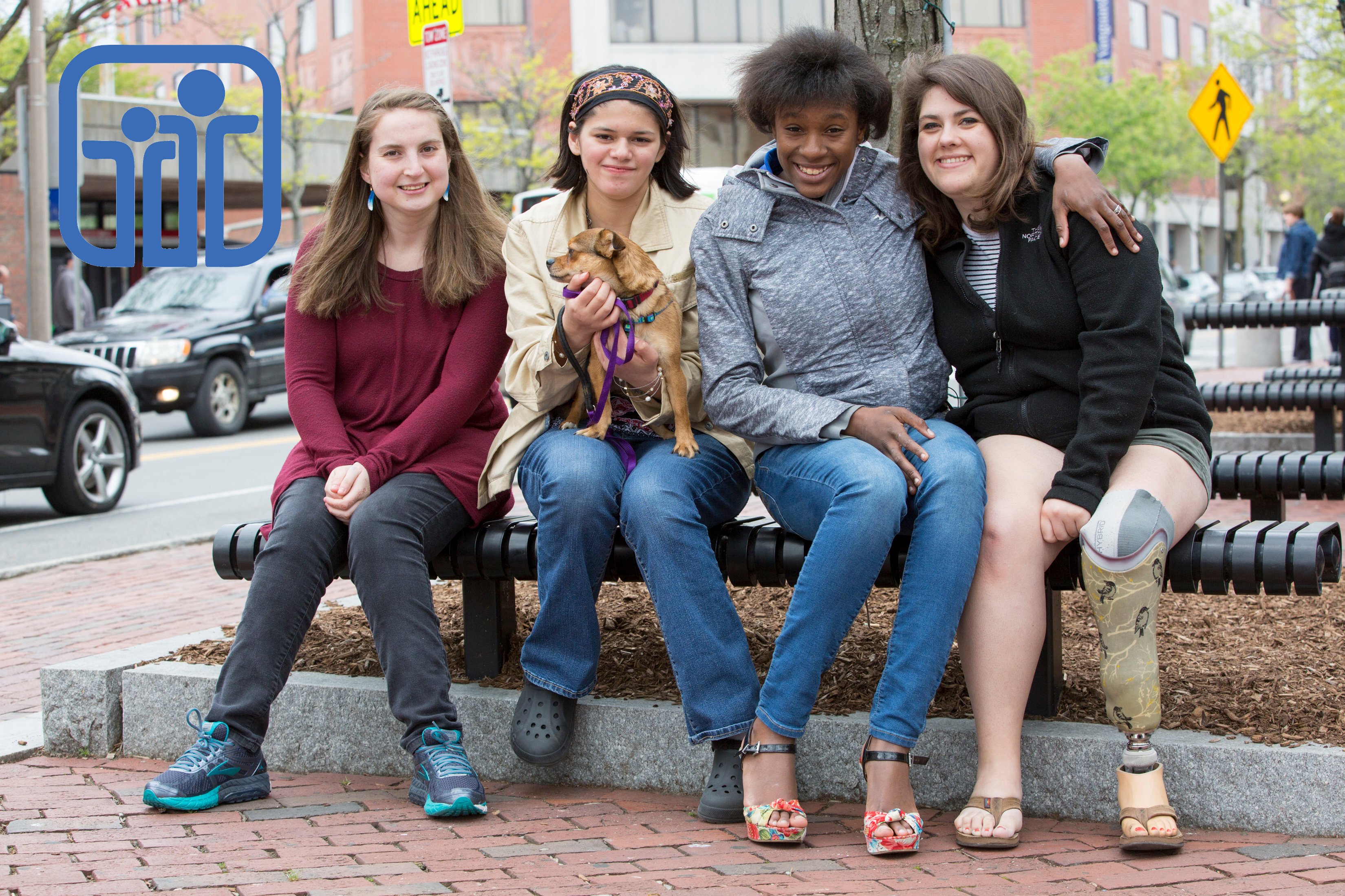 Four young women, one holding small dog, one with prosthetic leg, sit close together smilling on a sidewalk bench
