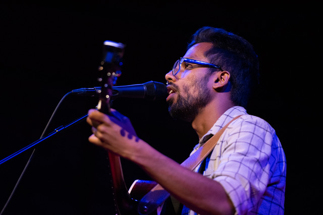 Prateek Poddar singing in to a microphone, holding a guitar.