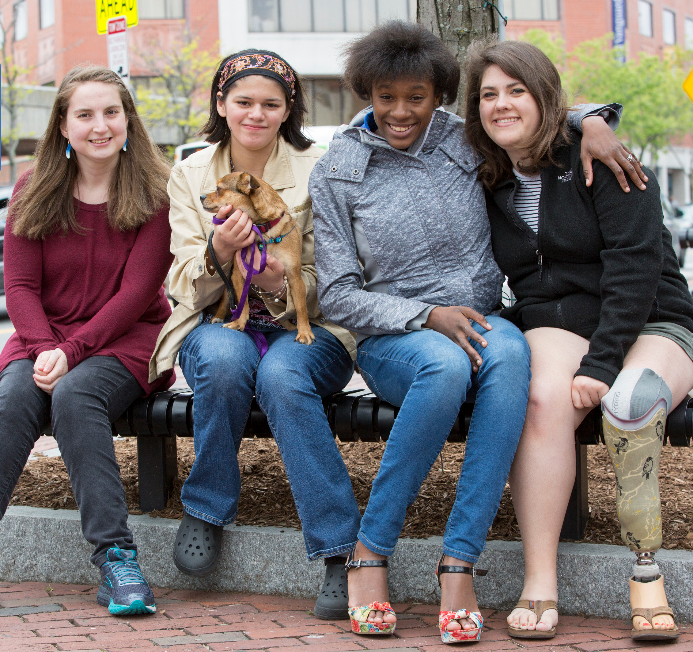Four young women sit together on outdoor bench, one holds small dog, one has prosthetic leg