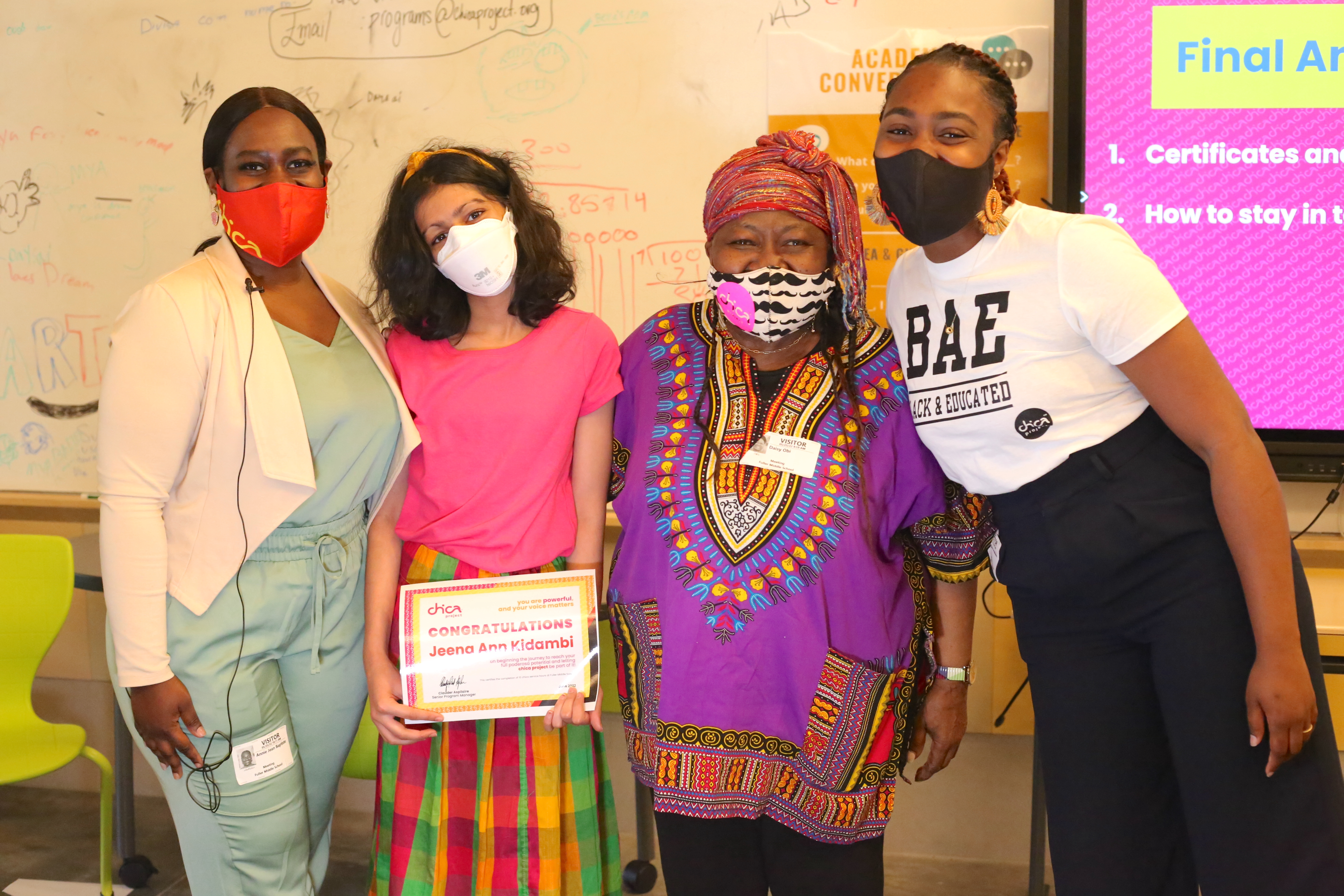 Four women in face masks and colorful clothes pose in front of classroom whiteboard
