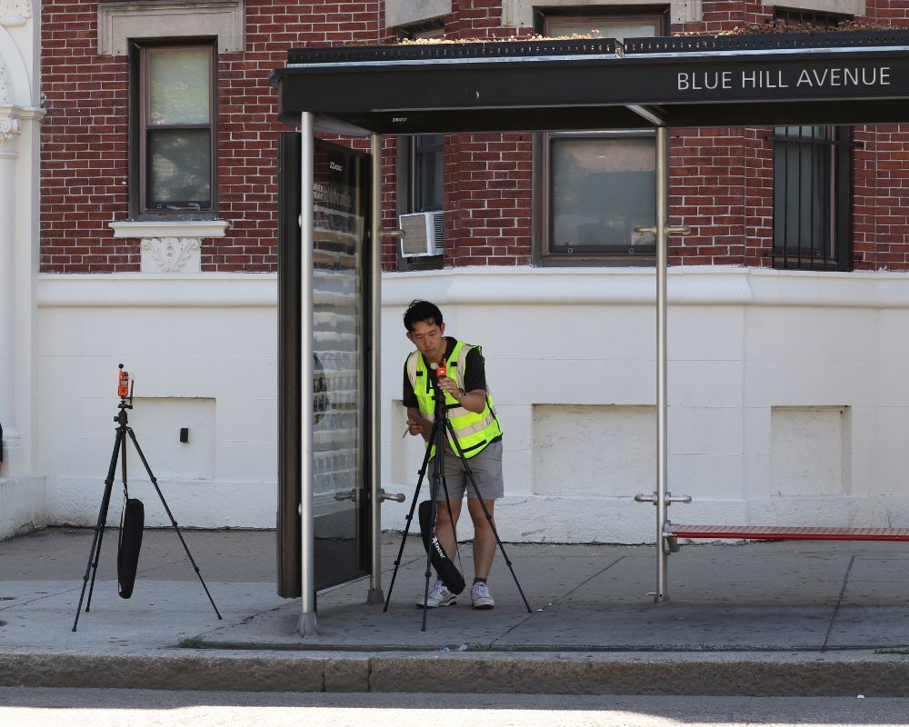 A man standing at a bus stop uses equipment on a tripod as part of the BCOOL data collection