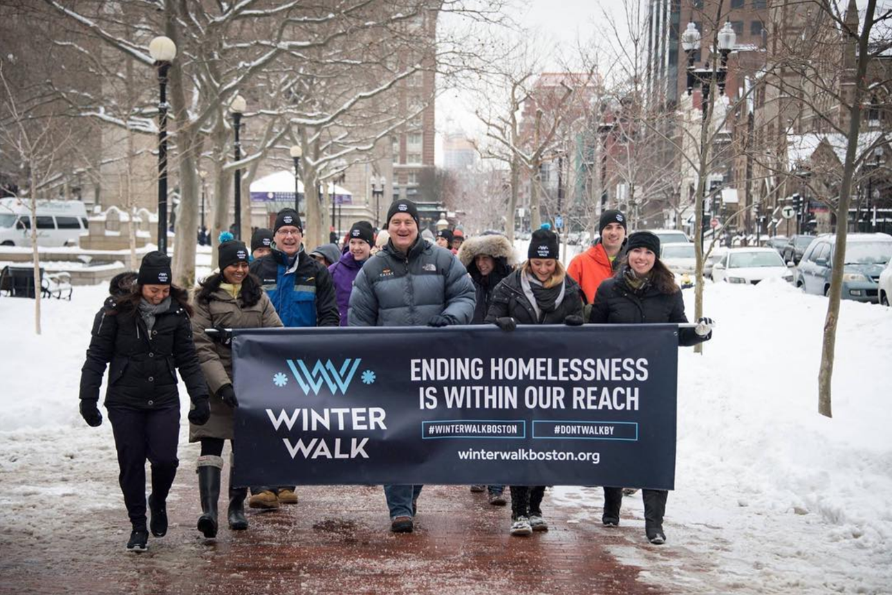A group of people march down a snowy sidewalk, they are holding a large banner that says "Winter walk, ending homelessness is within our reach."