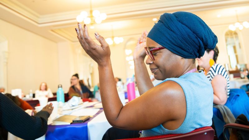 Sideview of woman in blue turban clapping with energy, seated at a table with others in background