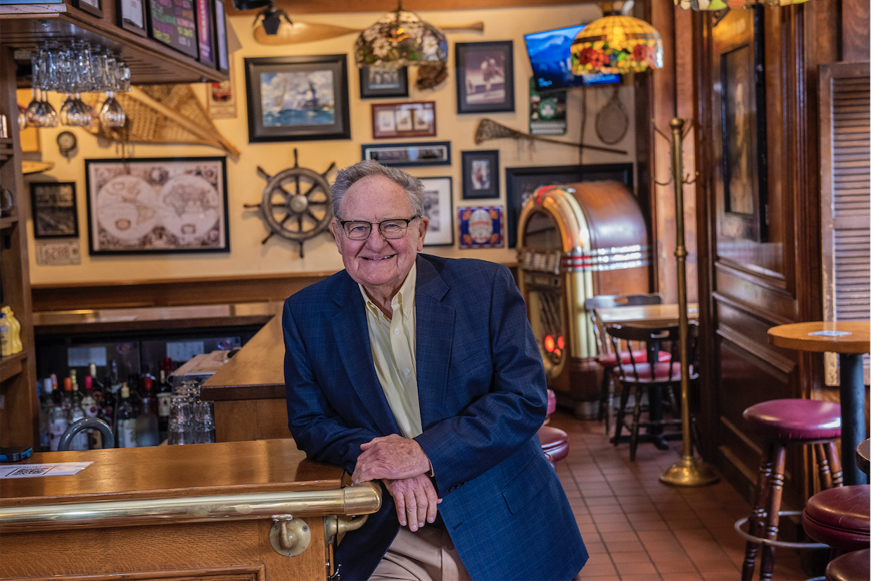 Tom Kershaw sitting at a bar. A yellow wall behind him is decorated with maps, photos, paintings, and a boat wheel.