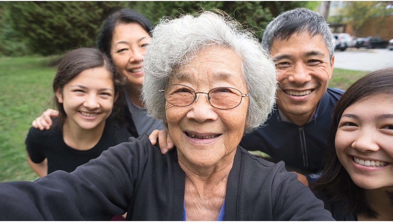 A selfie of an Asian family. An elderly woman in the center is taking the selfie. Behind her from left to right are a young girl, a woman, a man and another young girl. They are outside; green grass and trees are behind them. They're smiling and wearing black or navy blue shirts.