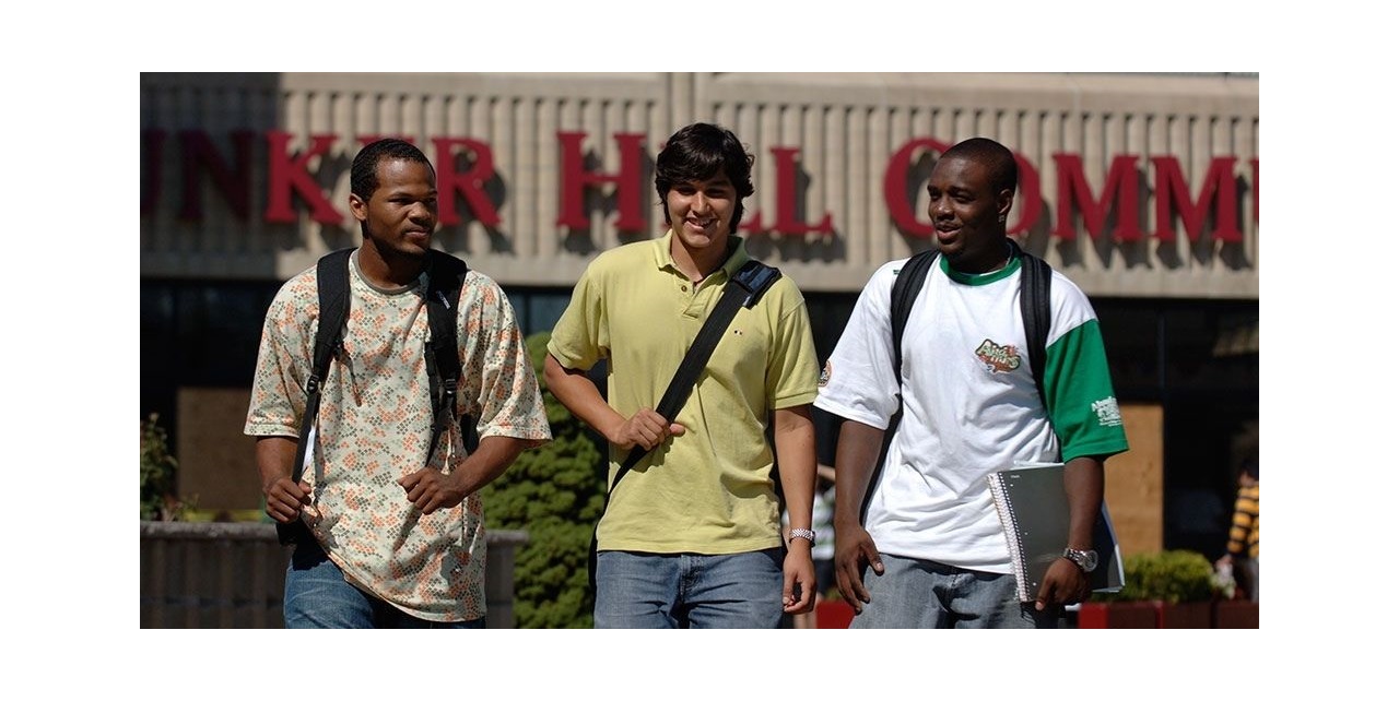 3 male students walking next to each other, smiling, with part of the main red Bunker Hill Community College sign on one of its buildings in the background.