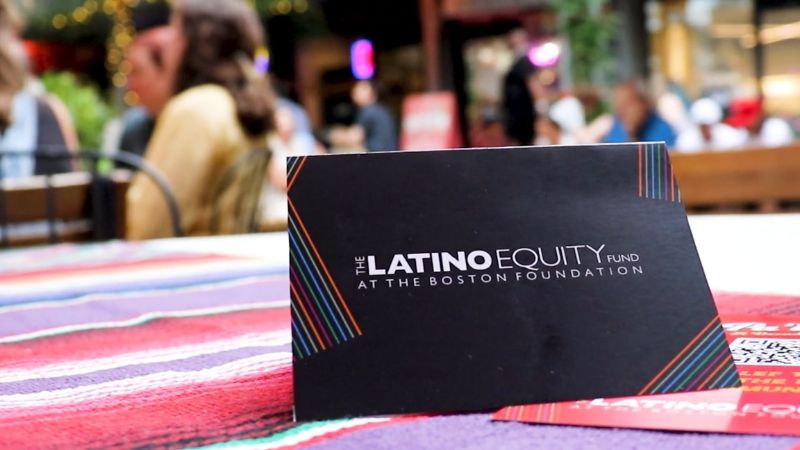 A card is standing up on a table at the Latino Equity Fund's 10 year celebration at Bow Market. The card is black and in white text reads the Latino Equity Fund at the Boston Foundation