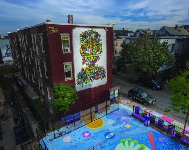 A brick building with a colorful mural on one side. A playground with colorfully painted pavement next to it. Blue skies at the top of the image with green trees surrounding the building/playground. 