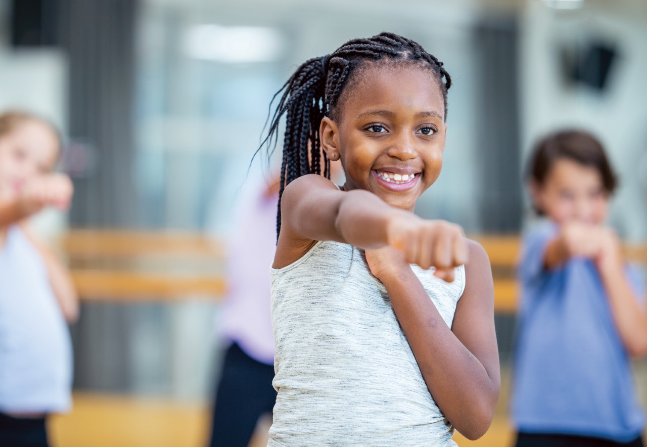 A young girl of color exercising, and behind her are other children doing the same.
