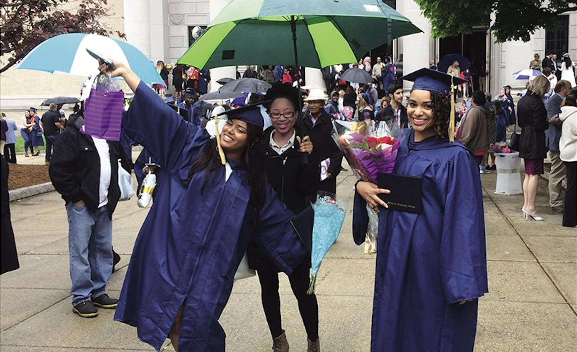 A picture of three women of color on graduation day. Two are wearing purple graduation robes, smiling, one of whom has her hands in the air. The third woman is standing in between the two graduates, wearing a black coat and holding an umbrella. Graduates and their families are in the background.
