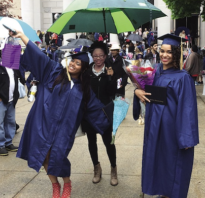 A picture of three women of color on graduation day. Two are wearing purple graduation robes, smiling, one of whom has her hands in the air. The third woman is standing in between the two graduates, wearing a black coat and holding an umbrella. Graduates and their families are in the background.
