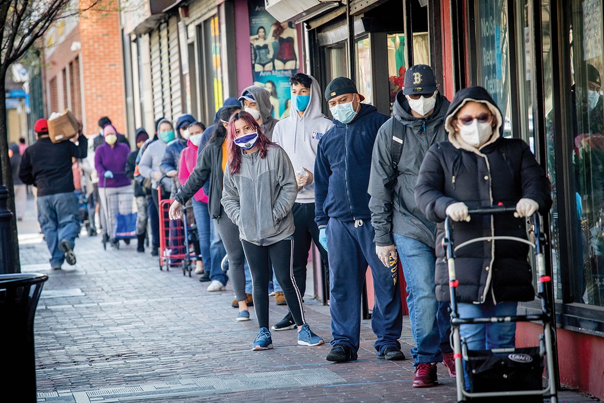A large number of people standing in line outside - all wearing masks - at a food bank.
