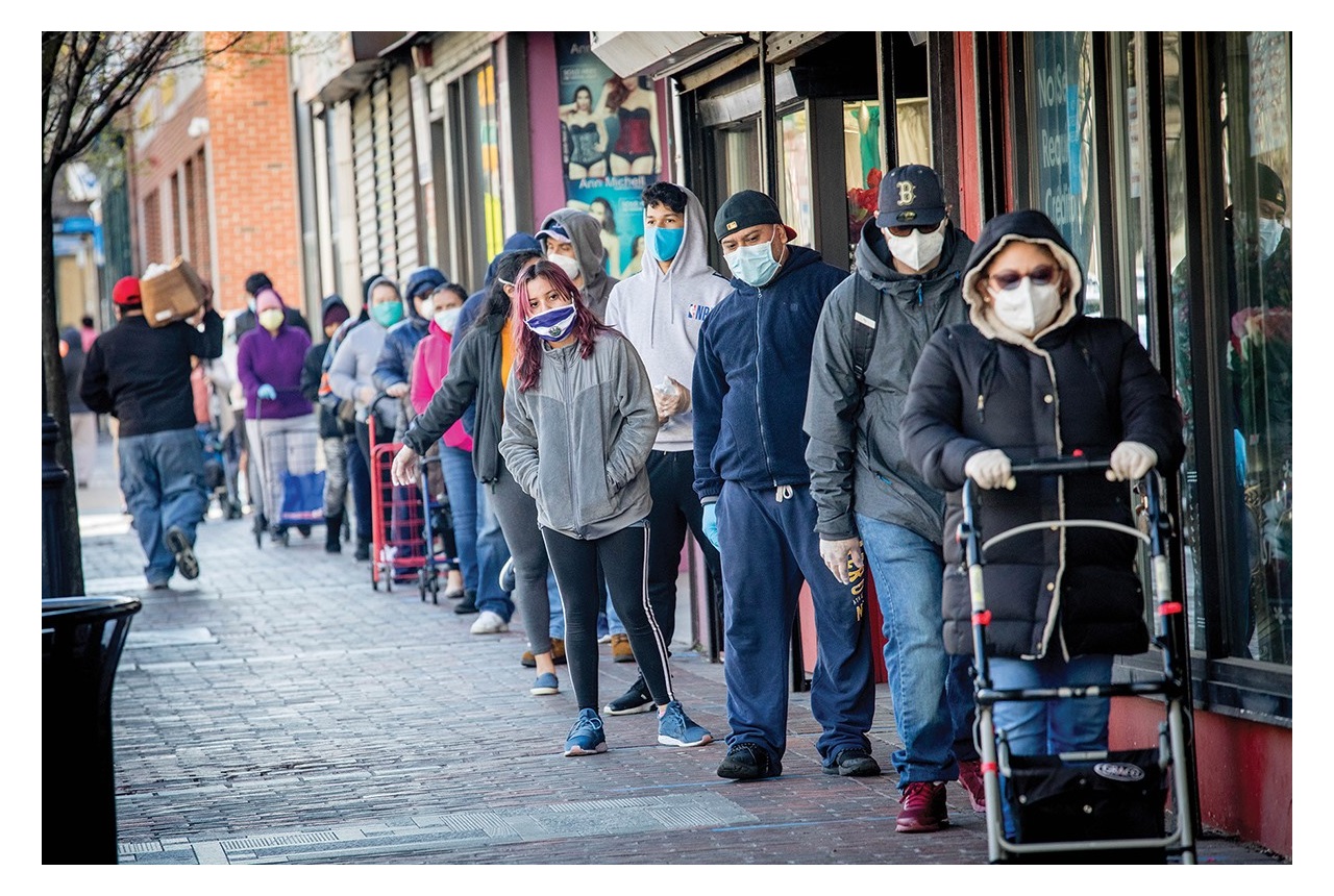A large number of people standing in line outside - all wearing masks - at a food bank.