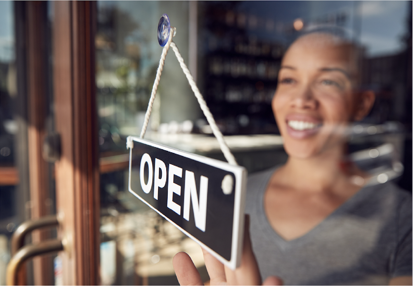 A woman of color standing behind a glass door, smiling as she hangs up an "open" sign.