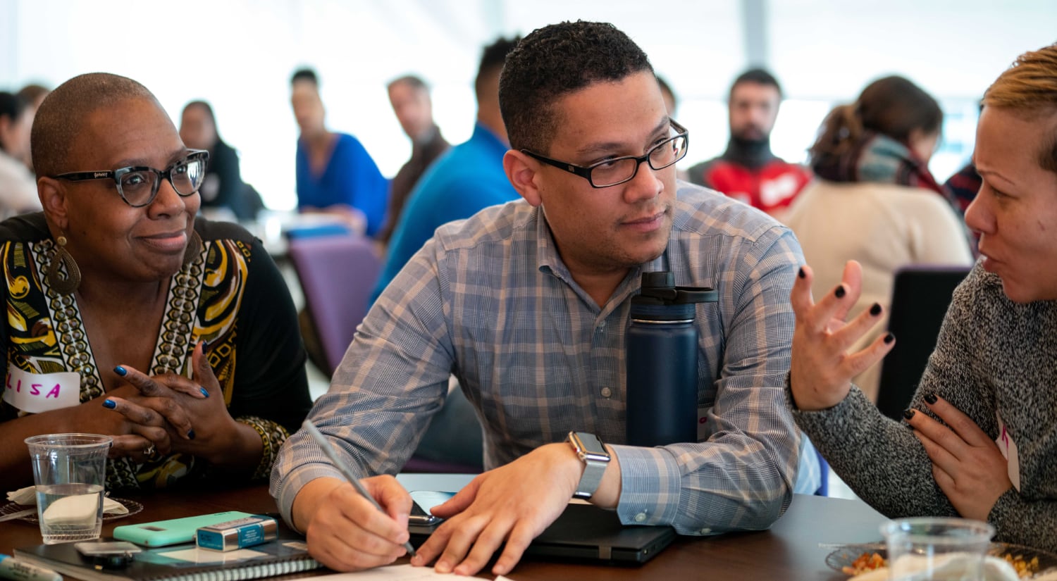A man sits between two women at a table. They are both talking to him, as he looks at the one to his left. He is holding a pencil to paper.